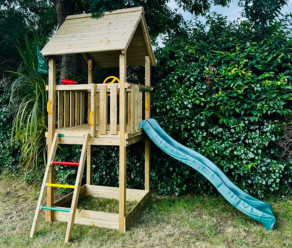 Compact wooden playhouse with green slide and climbing ladder placed next to a hedge